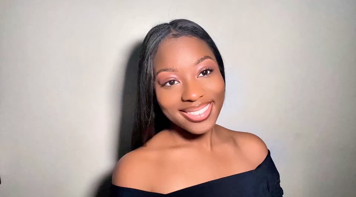 A smiling young Black woman with long dark hair wearing a black off-the-shoulder top.